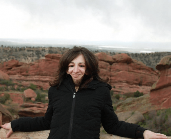 Picture of Alyse smiling, with dark brown long hair, wearing a black zip up jacket, standing outside in a grand canyon setting