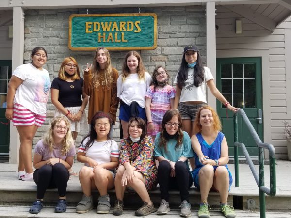 Picture of young teens smiling, outside in a camp setting. One row of kids is sitting down on steps while the second row is standing behind them. They are in front of a gray building that has a green and yellow sign that says Edwards Hall
