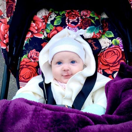 Picture of baby in a baby carrier decorated with rose background. The baby girl is wearing a white hat and coat, has blue eyes and is smiling. Also has a purple blanket