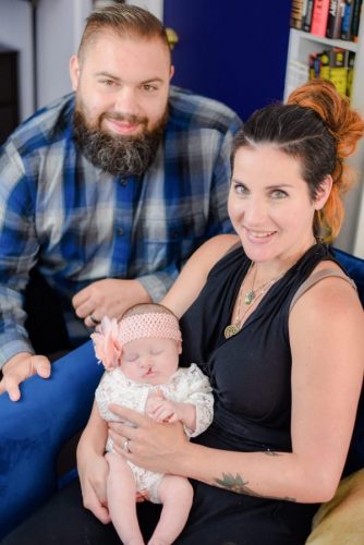 Picture of parents with a baby, Mother has long brown and blond hair, wearing a black dress with two green necklaces, she is holding a baby that is wearing a white onesie and a pink flower headband. The father has short brown hair, a brown and gray beard, and is wearing a blue plaid shirt