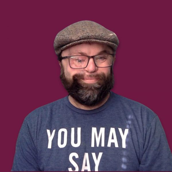 Picture of young man wearing a light gray newsboy cap, glasses, and a dark gray shirt that says You May Say. He has a dark brown and slightly gray beard and he is smiling