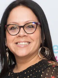 Picture of Lenica. A fair-skinned woman of color with straight black hair, large glasses with black frames, and wearing a black sgirt decorated with polka dots and flowers. She is smiling and is wearing large circular, dangling hoop earriings