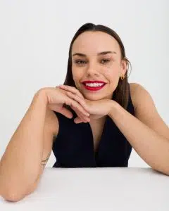 Picture of Phyllida Swift smiling wearing red lipstick, a sleeveless, v-neck, black blouse, and has straight dark brown hair. Her hands are being held together underneath her chin and her elbows are up on a white table.