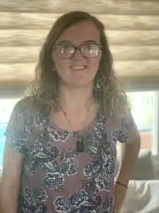 Emily Krebs (pictured) is smiling and has wavy, dark blonde, shoulder-length hair. She is wearing glasses, a necklace, and a pink shirt with blue designs on it