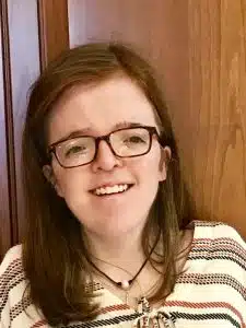 Morgan McGahan (pictured) is smiling and wearing black framed glasses, a white and brown striped shirt, and has shoulder length brown hair